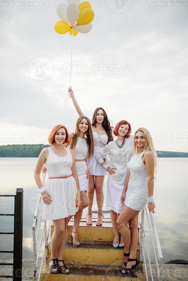 Five girls with balloons at hand weared on white dresses on hen party against pier on lake. photo