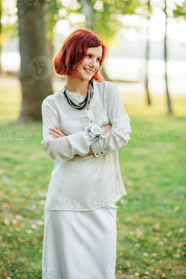 Portrait of red haired girl wearing on white dress against park at hen party. photo