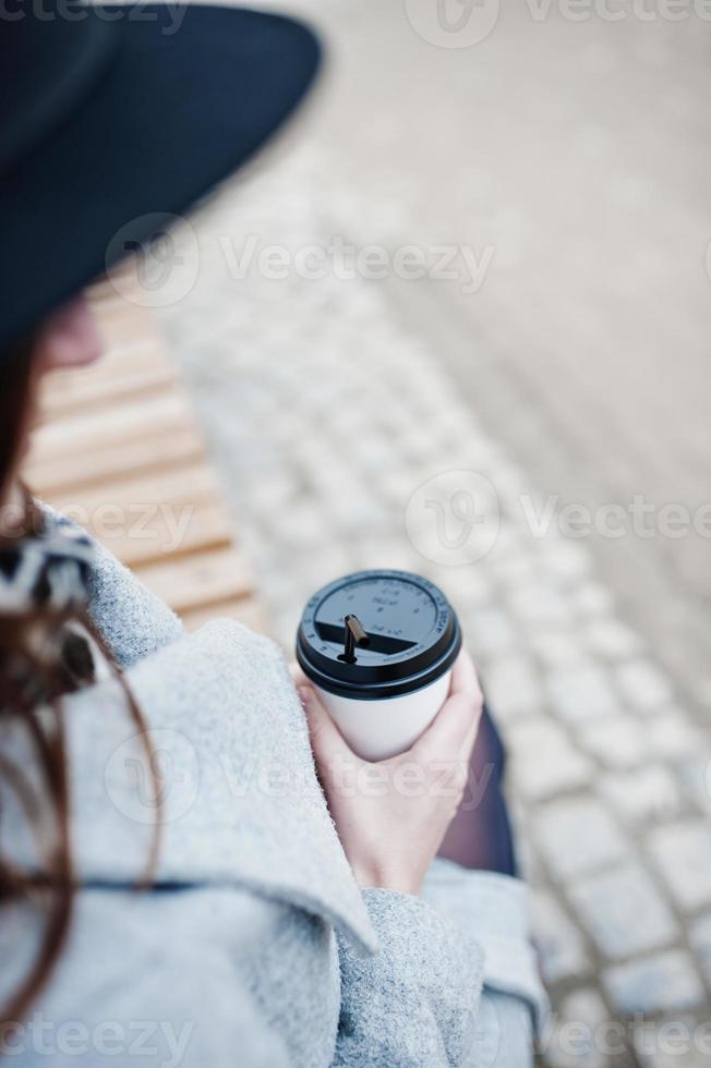 Close up plastic cup of coffee at hands of model girl in a gray coat and black hat sitting on bench at street of city. photo