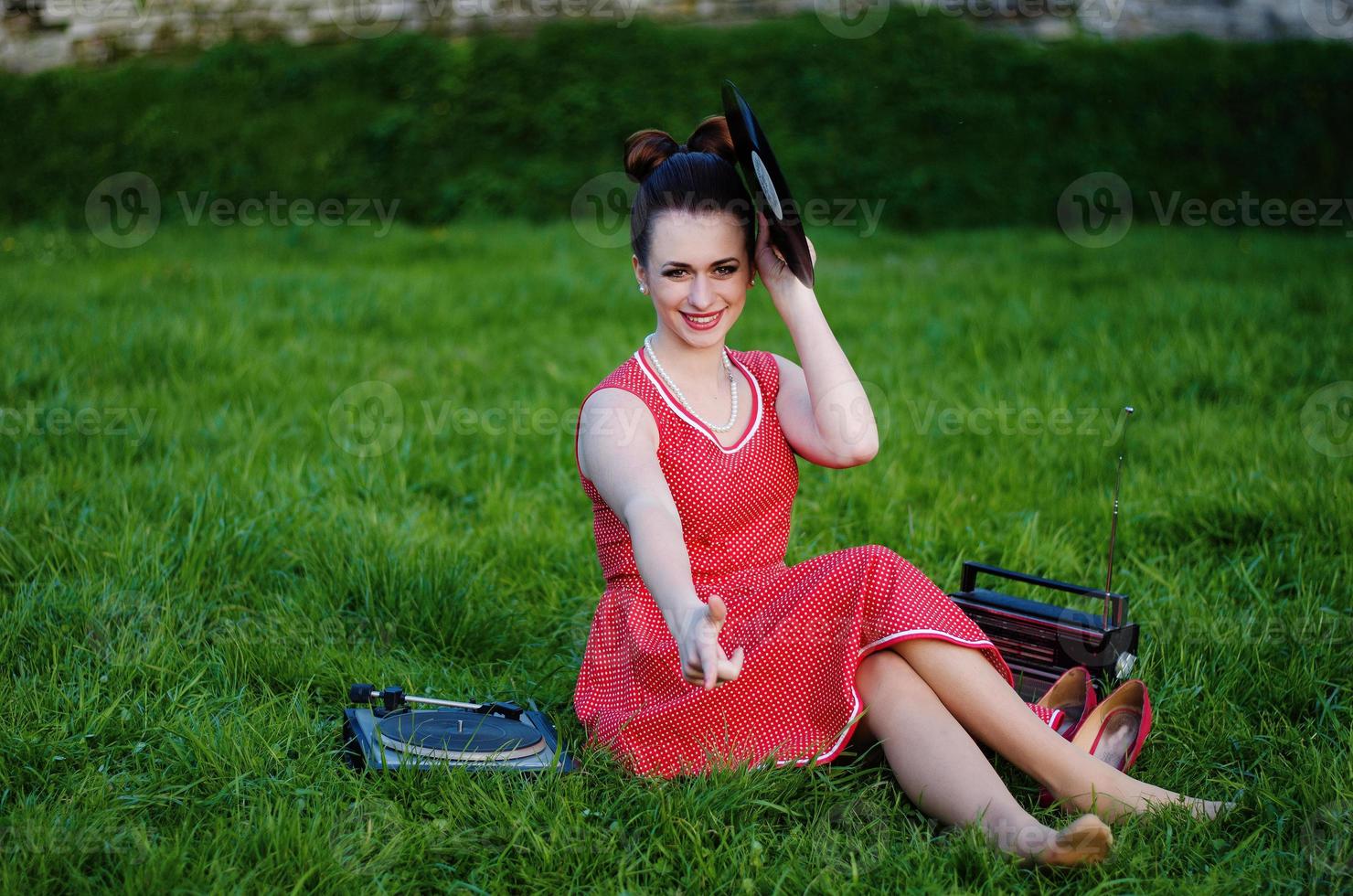 Portrait of young pinup girl wearing at retro vintage old-fashioned dress in peas sitting on grass wit retro radio and vinyl audio record in hands. photo