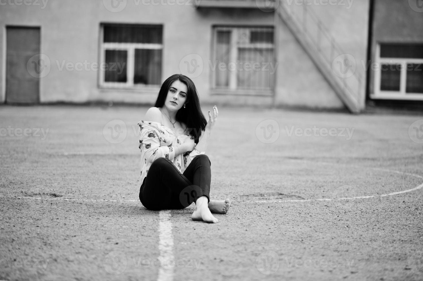 Young stylish teenage brunette girl on shirt, pants and high heels shoes, sitting on pavement and posed background school backyard. Street fashion model concept. photo