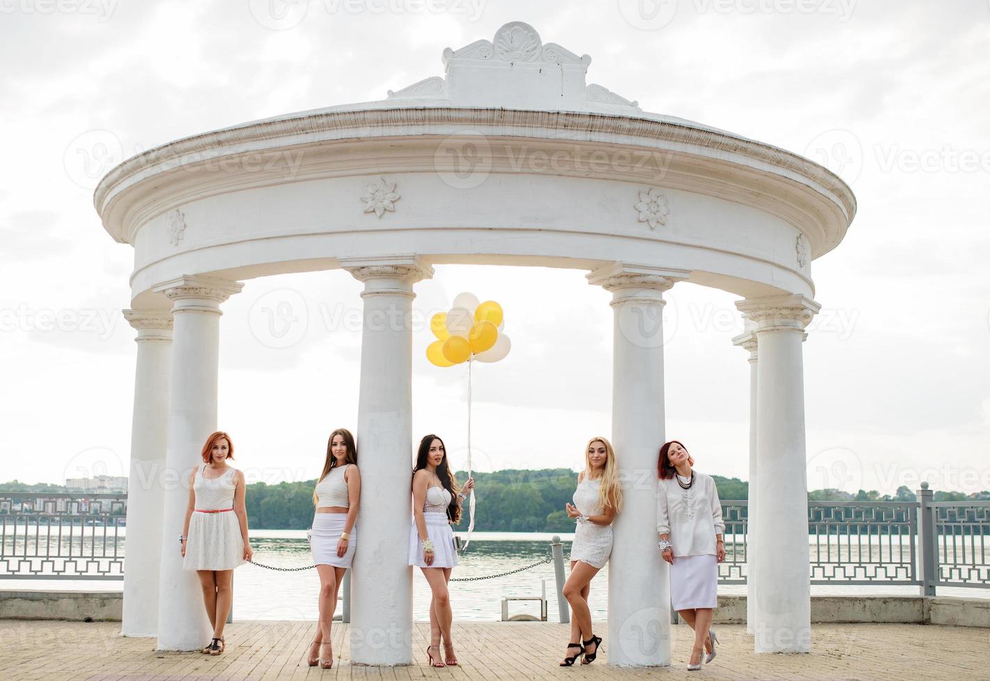 Five girls with balloons at hand weared on white dresses on hen party against white columns of arch. photo