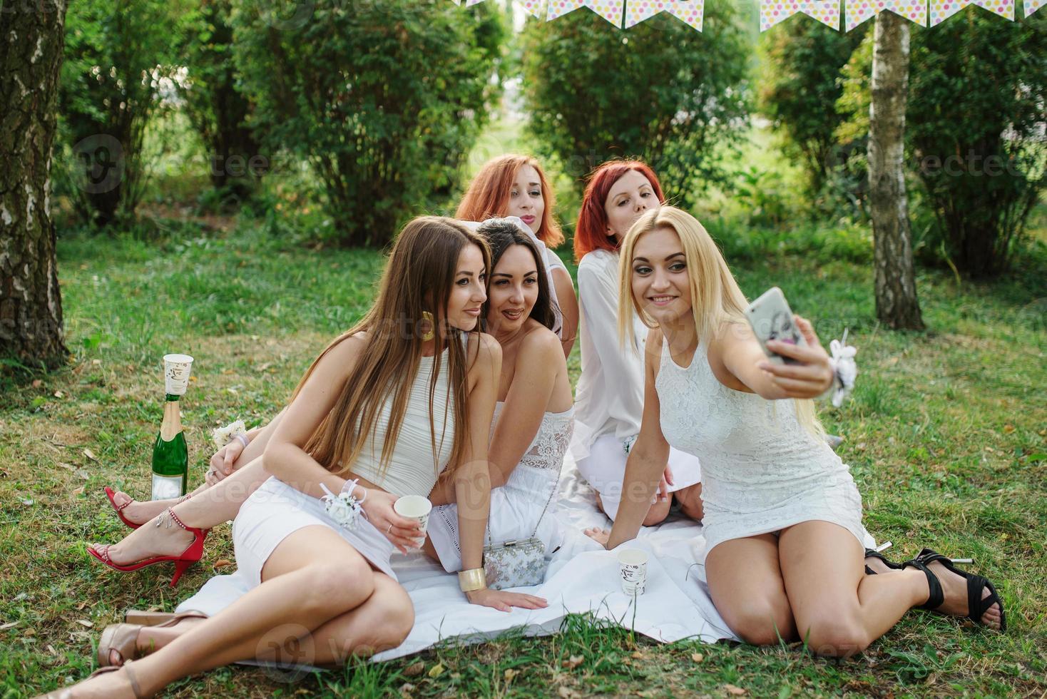 cinco chicas vestidas de blanco en una despedida de soltera sentadas al aire libre y haciéndose selfie en el parque. foto