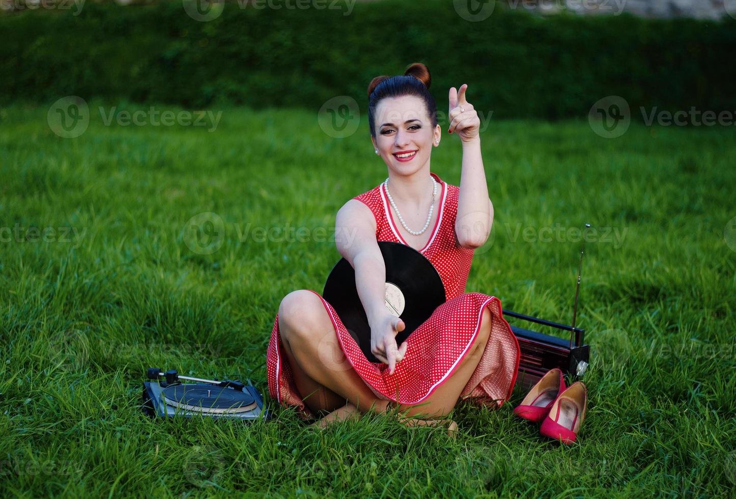 Portrait of young pinup girl wearing at retro vintage old-fashioned dress in peas sitting on grass wit retro radio and vinyl audio record in hands. photo