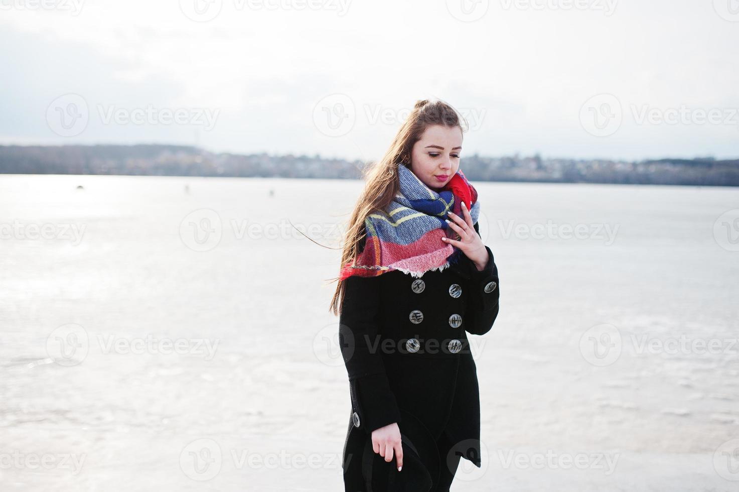 chica joven casual en abrigo negro, bufanda y sombrero contra el río congelado en el clima soleado de invierno. foto