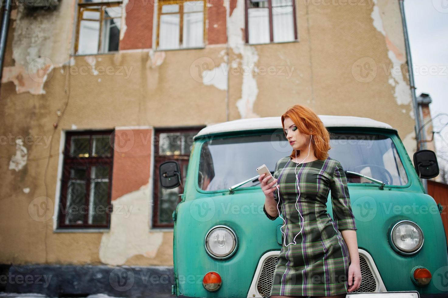 Young red haired girl lady with mobile cell phone and headphones, wearing on checked dress background old vintage turqoise minivan bus. photo