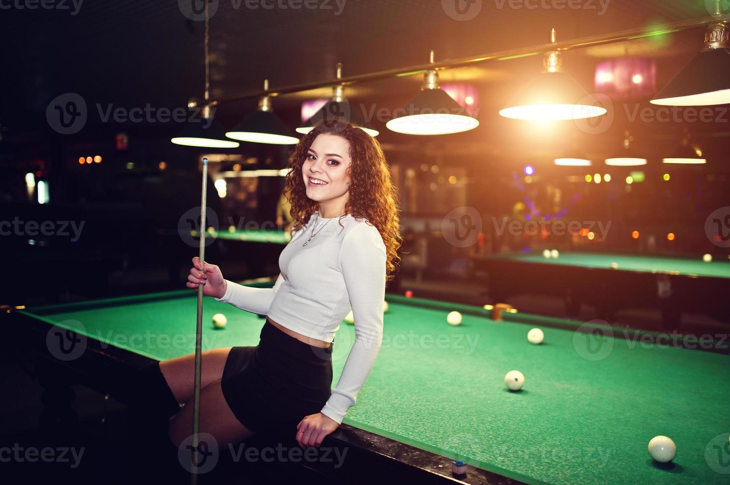 Young curly girl posed near billiard table. Sexy model at black mini mini skirt play russian snooker. Play game and fun concept. photo