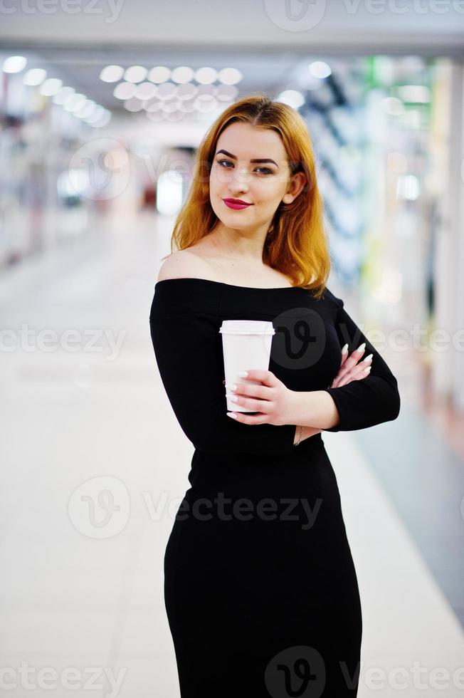 Fashion red haired girl wear on black dress with bright make holding coffee cup at trade shopping center. Photo toned style Instagram filters.