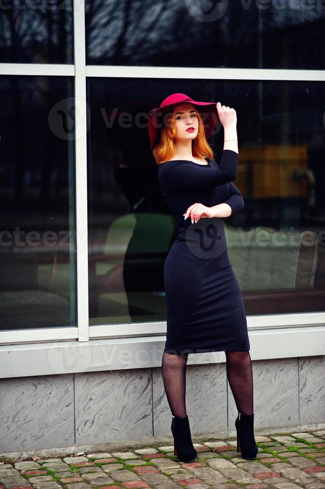 Portrait of fashion red haired girl on red hat and black dress with bright make up posed against large window. Photo toned style Instagram filters.