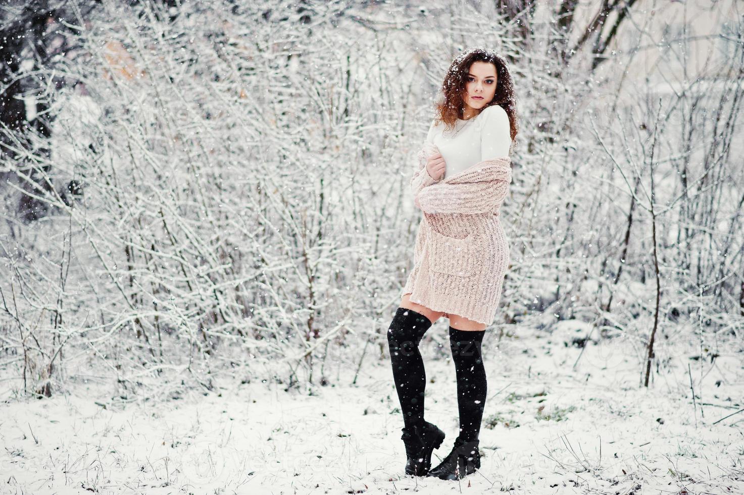 Curly brunette girl background falling snow, wear on warm knitted sweater, black mini skirt and wool stockings. Model on winter. Fashion portrait at snowy weather. Instagram toned photo. photo