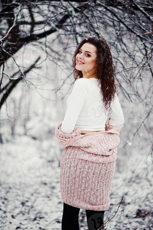 Back of curly brunette girl background falling snow, wear on warm knitted sweater, black mini skirt and wool stockings. Model on winter. Fashion portrait at snowy weather. Instagram toned photo. photo