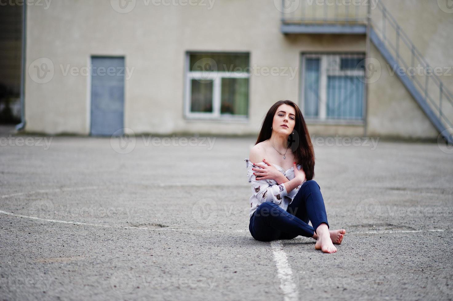 joven morena adolescente elegante con camisa, pantalones y zapatos de tacones altos, sentada en el pavimento y posando en el patio trasero de la escuela. concepto de modelo de moda callejera. foto