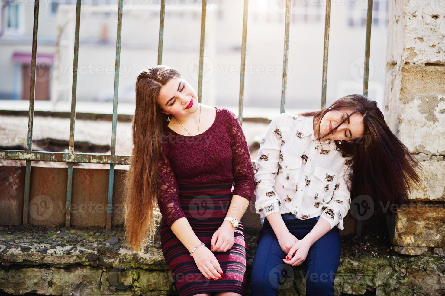 Two young teenages girl sitting against iron fence and moving with they hair. Plump girl vs thin. Friends teen. photo