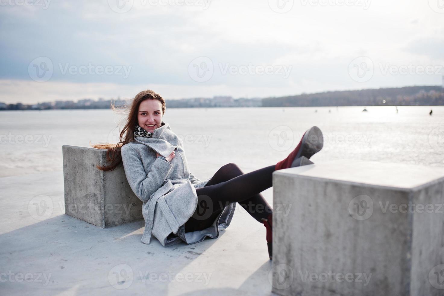 Young model girl in gray coat posed sitting near stone cube against freeze lake. photo