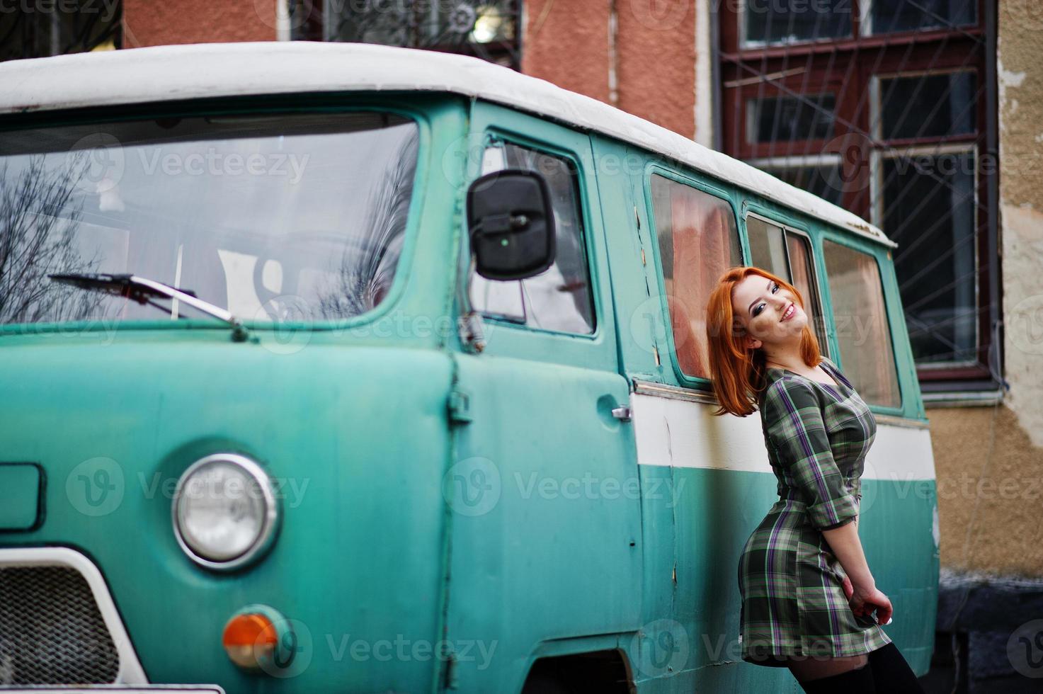 Young red haired girl posed on checkered dress background old retro cyan minivan. photo