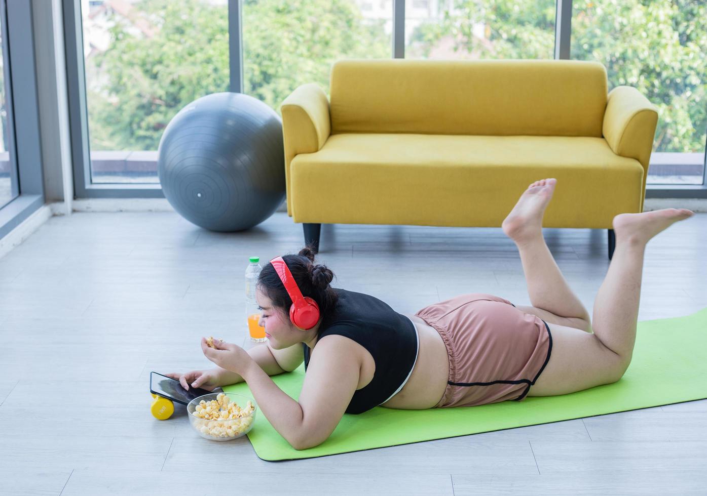 mujer asiática con sobrepeso usando tableta y. usando auriculares para escuchar música y ella está haciendo ejercicio en casa, una chica asiática disfruta comiendo comida y palomitas de maíz foto