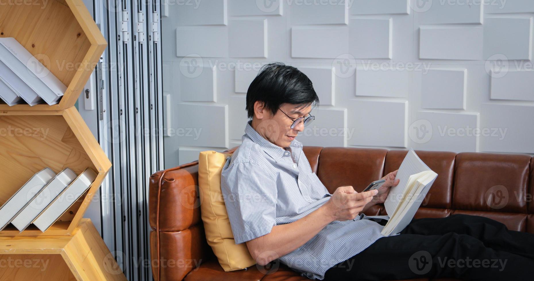 Senior Asian man reading book and using smart phone on sofa in living room at home  Portrait of Asian elderly man is Relaxing and Happiness With Read a Magazine photo