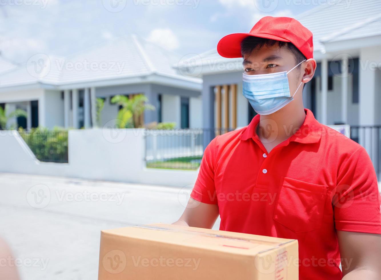 repartidores asiáticos con uniforme rojo con gorra roja y máscara facial manejando cajas de cartón para dárselas a la clienta frente a la casa. compras en línea y entrega urgente foto