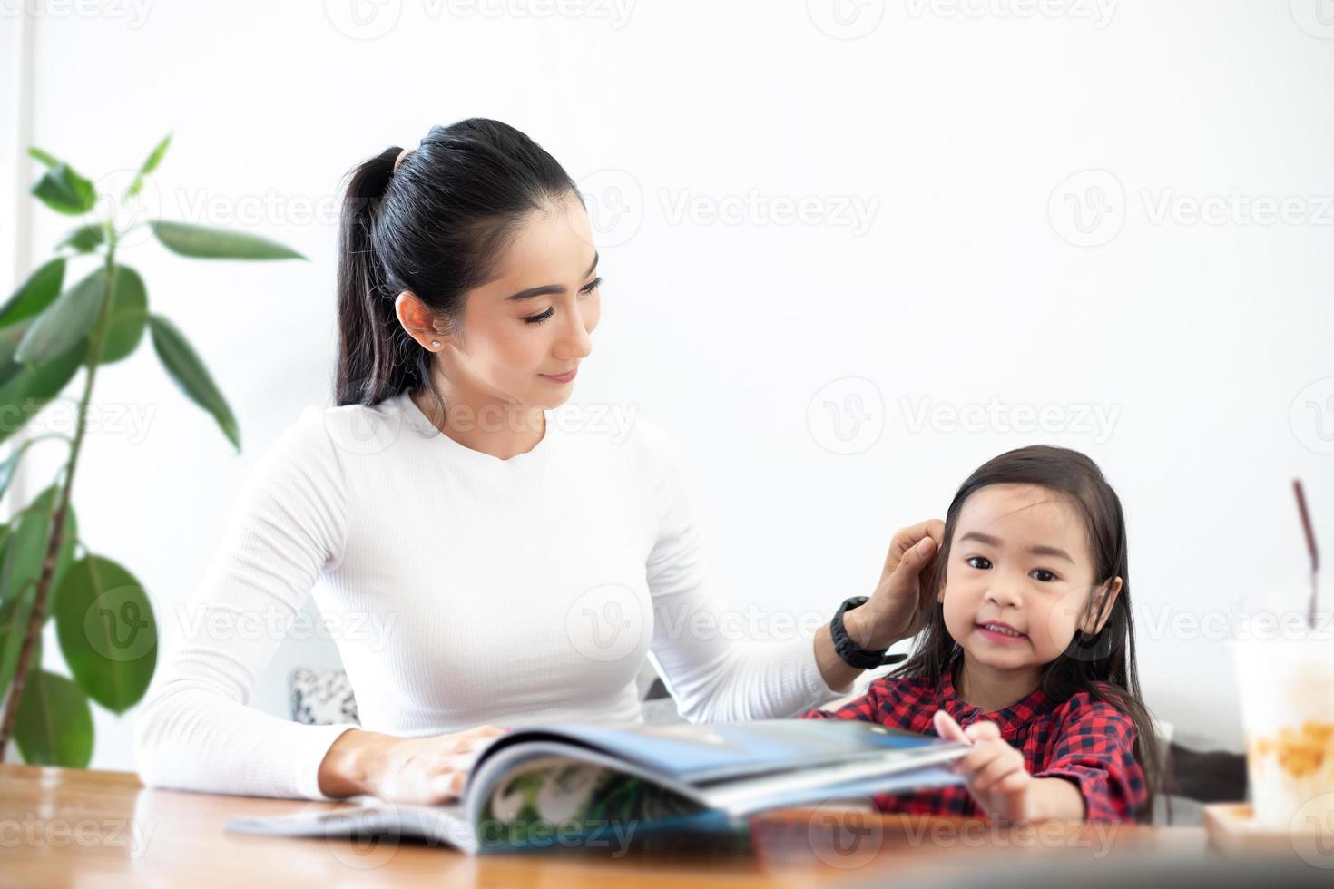 An Asian mother is teaching her daughter to read a book during the semester break on the living table and having cold milk on the table at home. Educational concepts and activities of the family photo