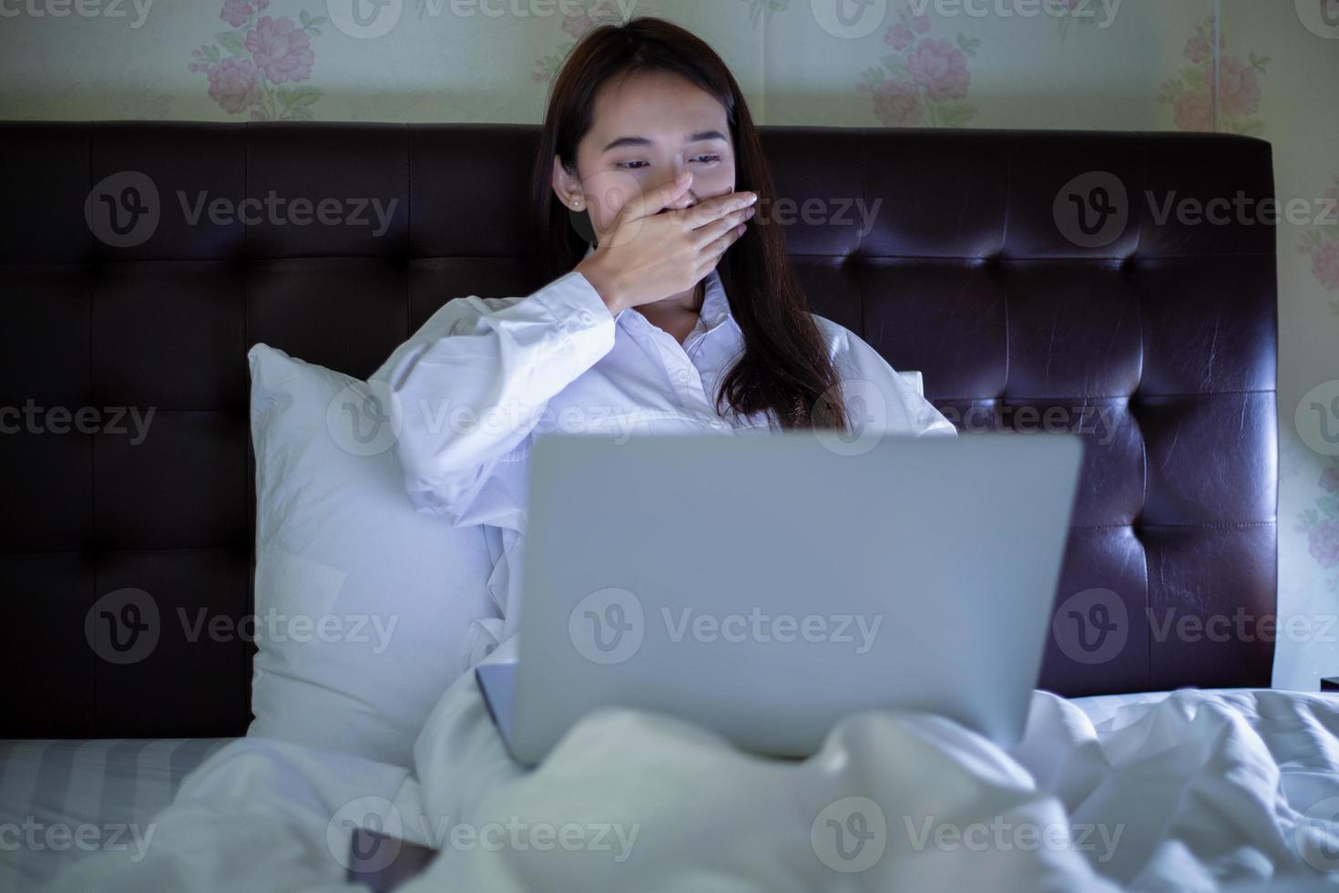 mujer asiática que usa un cuaderno para trabajar de noche en la cama y bosteza y tiene sueño. foto