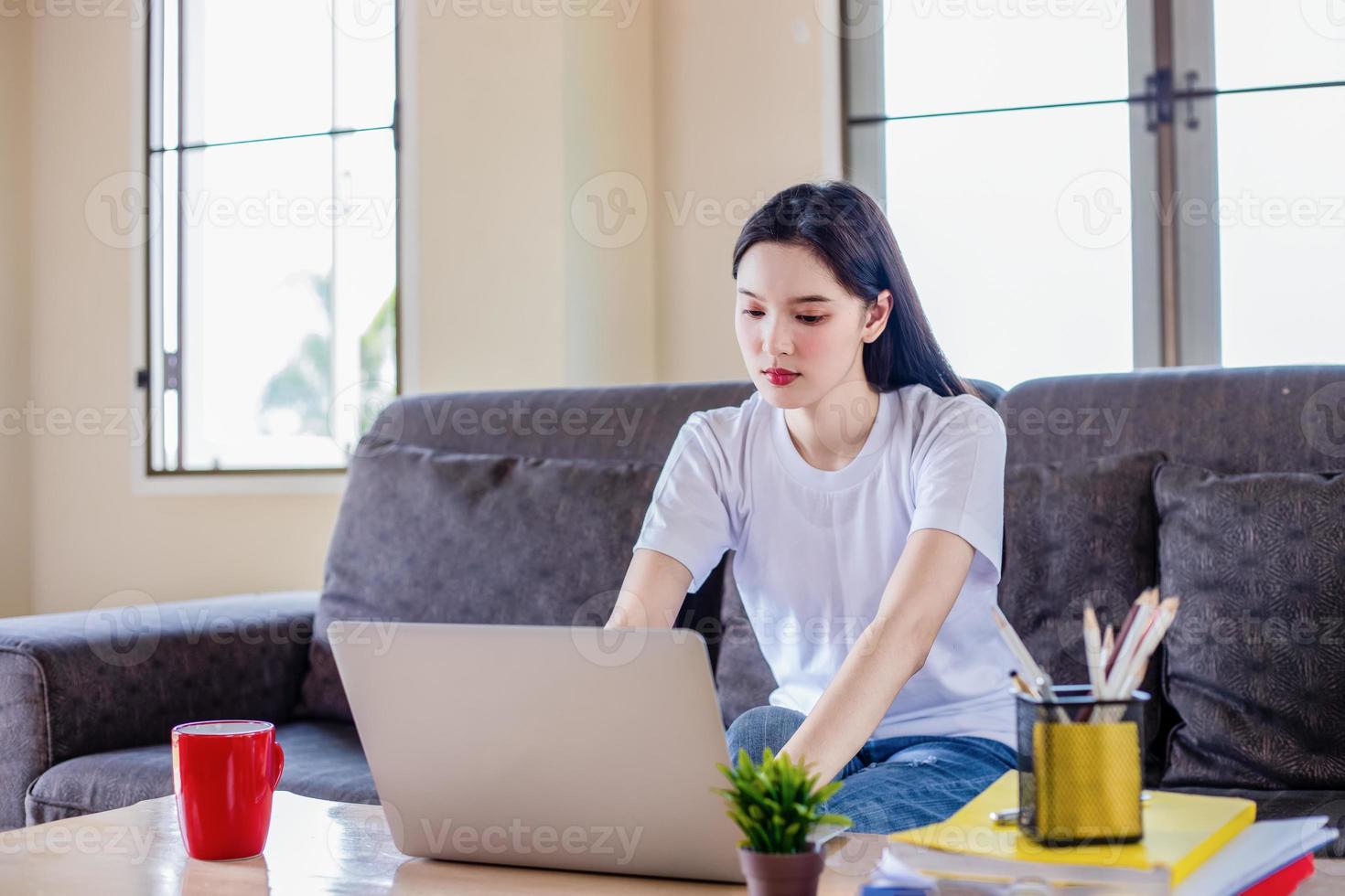 Asian woman pleased happy cheerful cute beautiful is using a notebook computer and sitting on sofa for working from home. She is quarantined at home during the coronavirus epidemic. photo