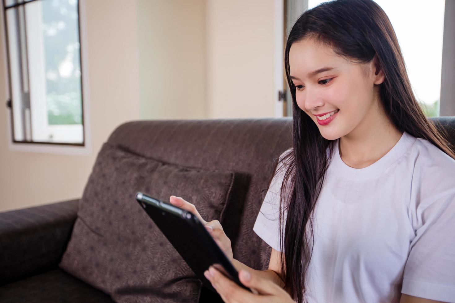 Asian young woman happy cheerful cute beautiful is playing digital pad tablet computer and sitting on the sofa at home, smiling lady using app reading electronic book relax in living room photo