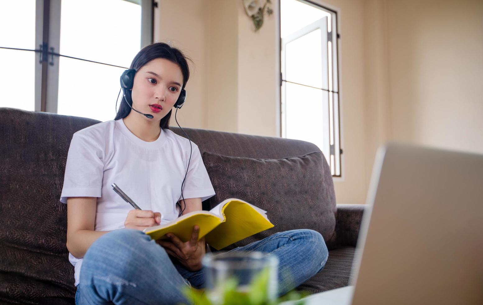 una estudiante asiática feliz usa audífonos estudia en línea viendo un podcast de seminario web en una computadora portátil escuchando un curso de educación llamada en conferencia y escribe notas mira la computadora portátil sentada en un sofá en casa foto