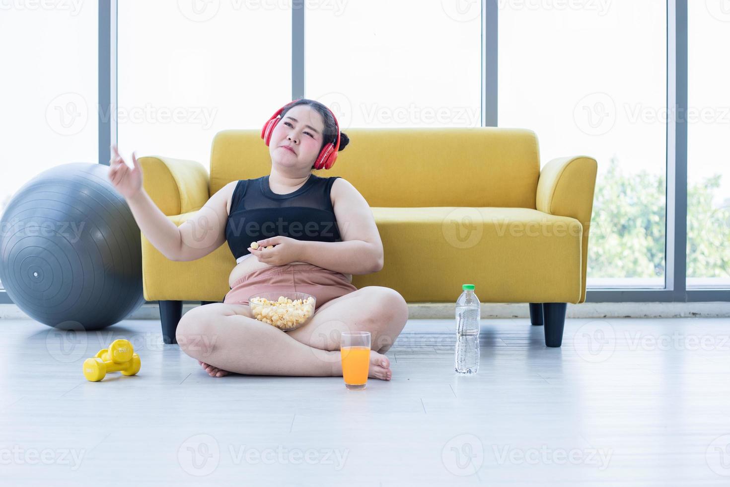 mujer asiática con sobrepeso usando tableta y. usando auriculares para escuchar música y ella está haciendo ejercicio en casa, una chica asiática disfruta comiendo comida y palomitas de maíz foto