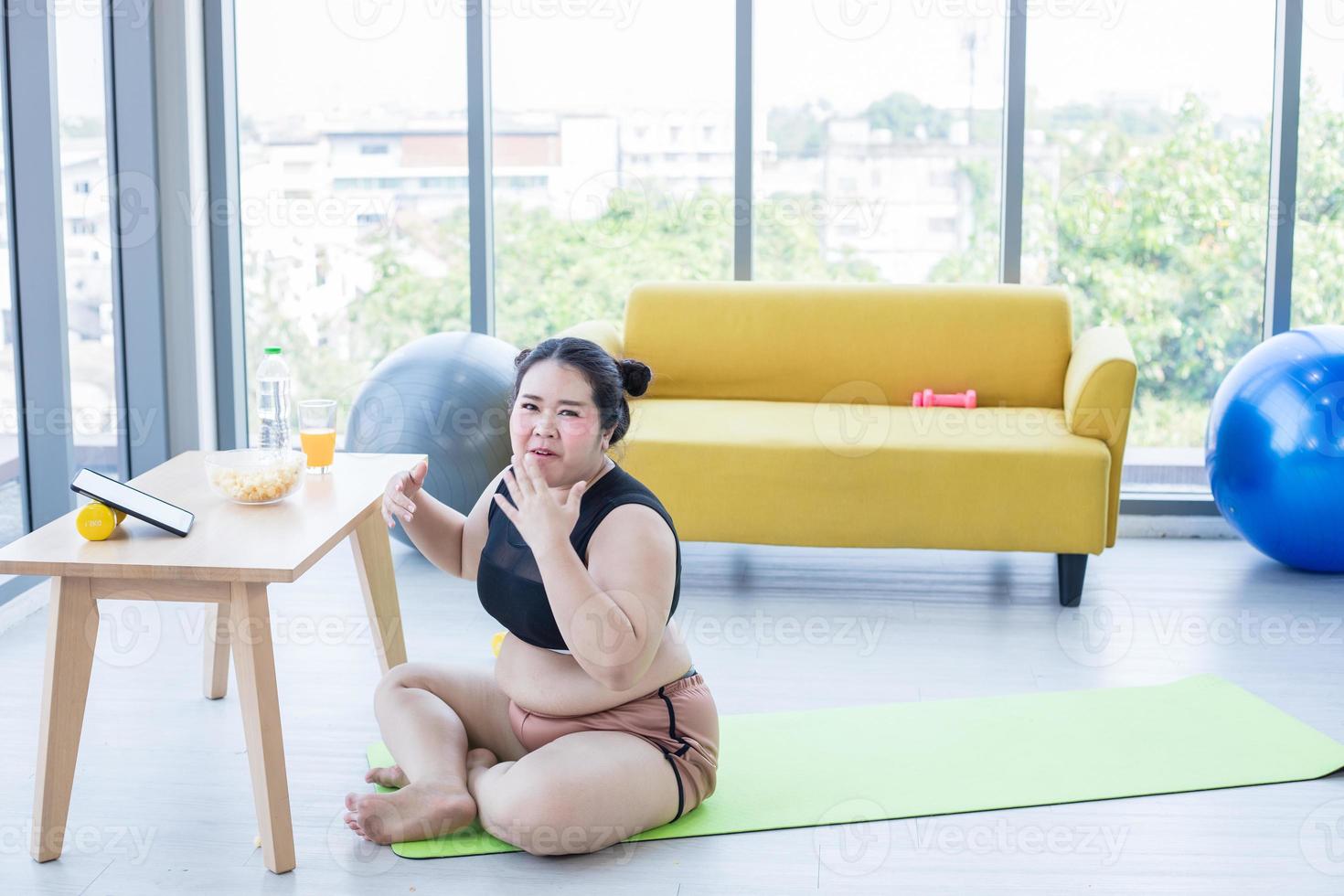 mujer asiática con sobrepeso usando tableta y. usando auriculares para escuchar música y ella está haciendo ejercicio en casa, una chica asiática disfruta comiendo comida y palomitas de maíz foto