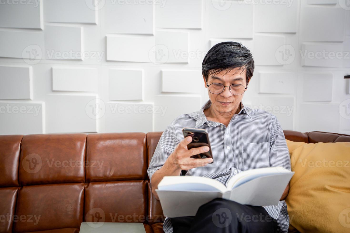 Senior Asian man reading book and using smart phone on sofa in living room at home  Portrait of Asian elderly man is Relaxing and Happiness With Read a Magazine photo