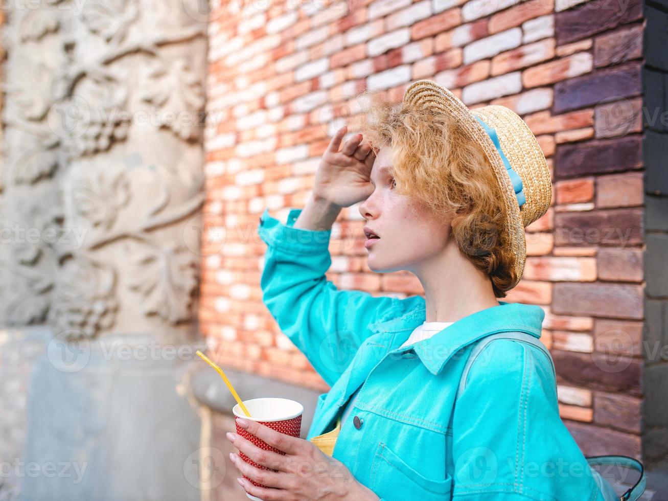 young cheerful curly redhead woman in blue denim jacket and straw hat holding cup of coffee and surprised by landmarks. Fun, summer, travel, fashion, youth concept photo