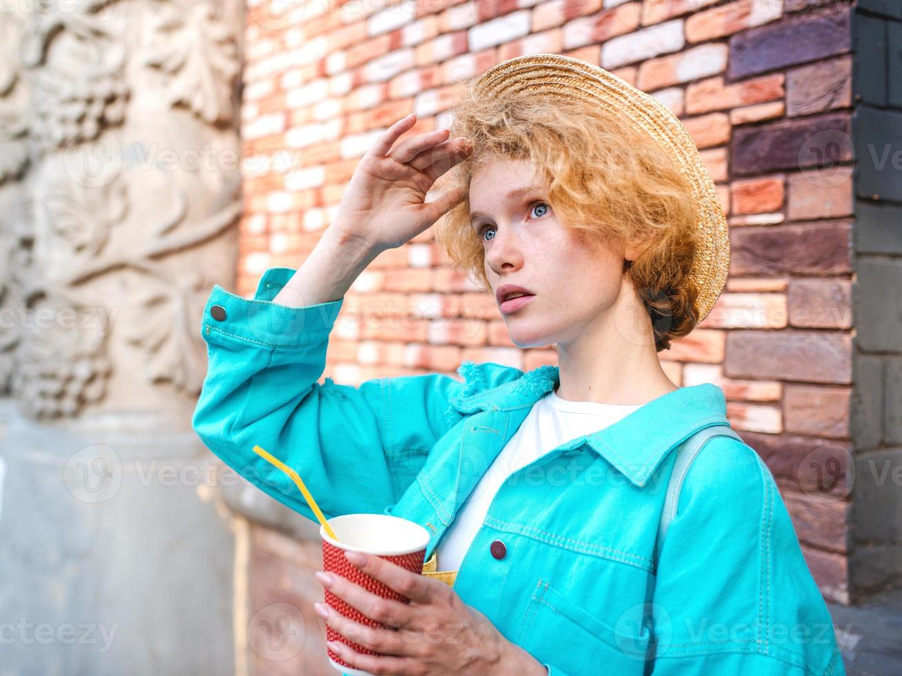 young cheerful curly redhead woman in blue denim jacket and straw hat holding cup of coffee and surprised by landmarks. Fun, summer, travel, fashion, youth concept photo