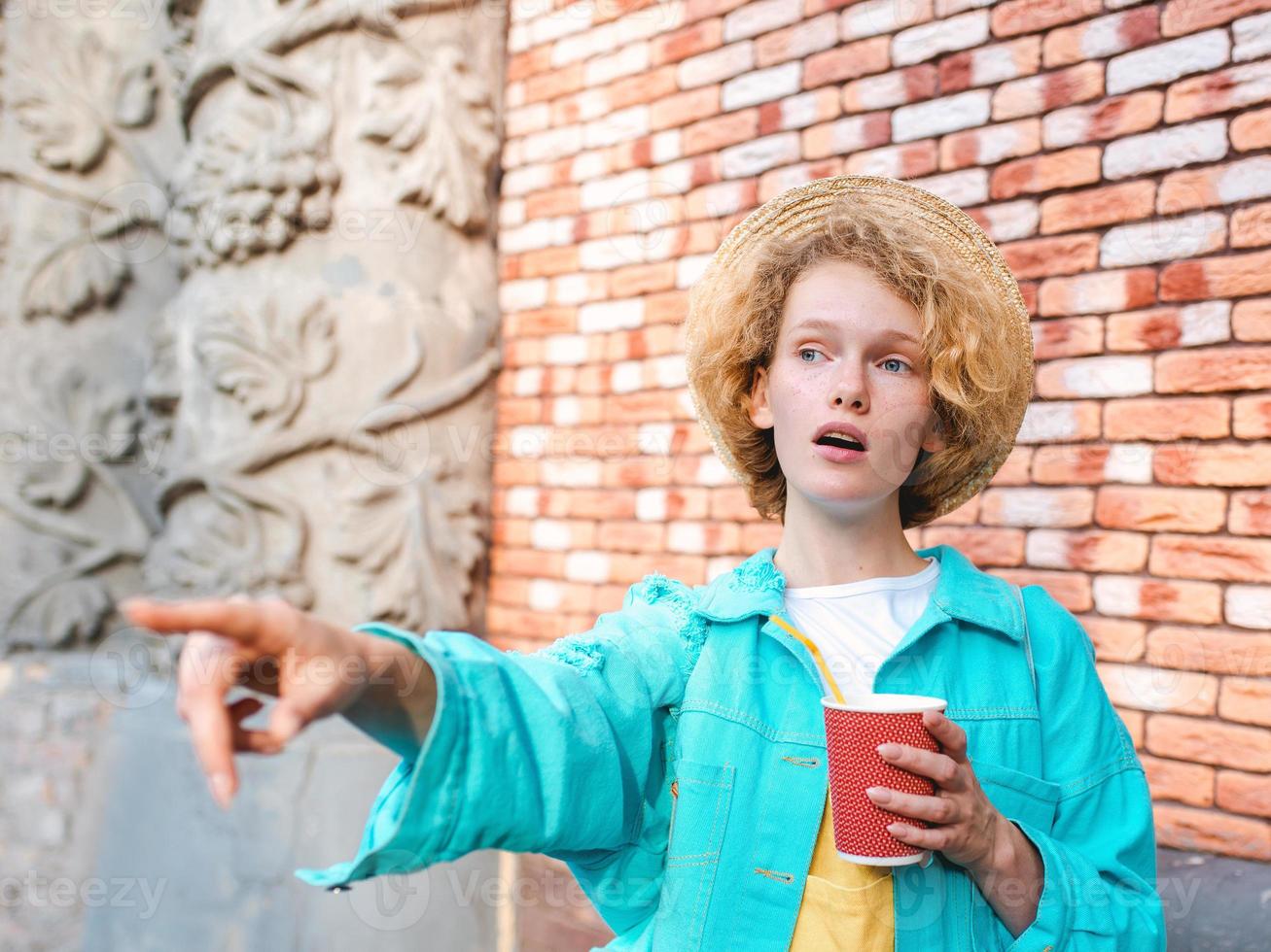 young cheerful curly redhead woman in blue denim jacket and straw hat holding cup of coffee and surprised by landmarks. Fun, summer, travel, fashion, youth concept photo