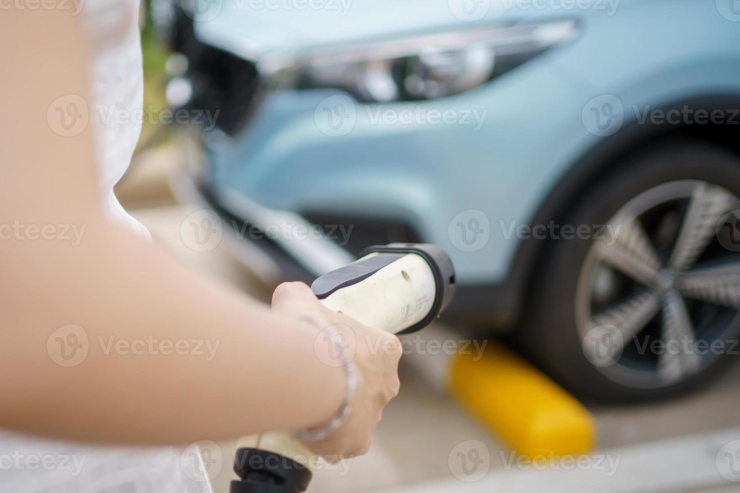 feliz mujer asiática sosteniendo un conector de carga dc - ccs tipo 2 ev en la estación de carga ev, mujer preparando un ev - conector de carga de vehículos eléctricos para recargar un vehículo. foto