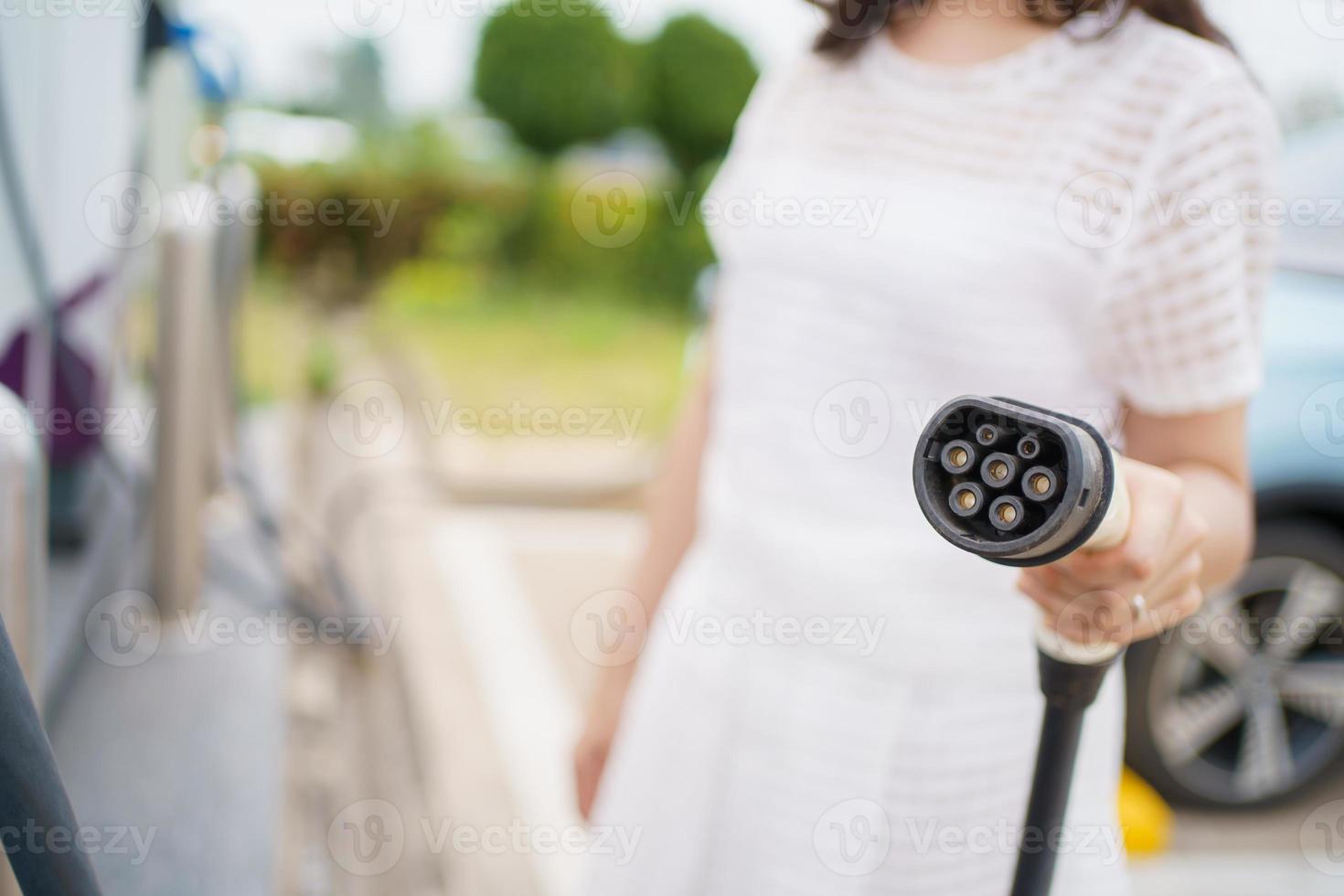 Happy Asian woman holding a DC - CCS type 2 EV charging connector at EV charging station, woman preparing an EV - electric vehicle charging connector for recharge a vehicle. photo
