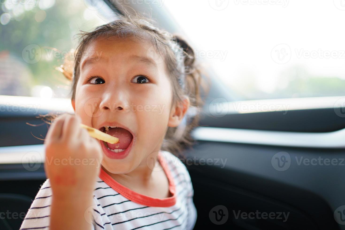 pequeña niña asiática que se queda en el auto y come papas fritas, una chica encantadora disfruta comiendo bocadillos mientras viaja en el auto. foto