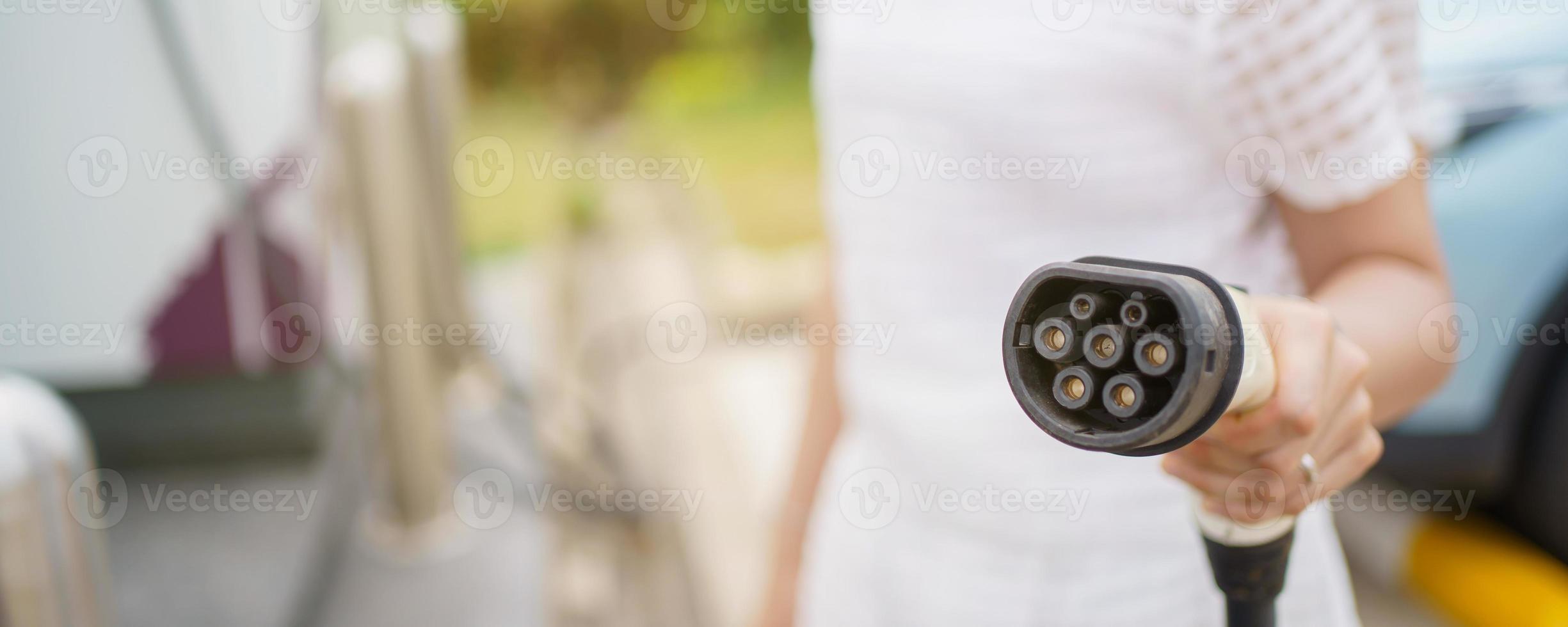 Happy Asian woman holding a DC - CCS type 2 EV charging connector at EV charging station, woman preparing an EV - electric vehicle charging connector for recharge a vehicle. photo