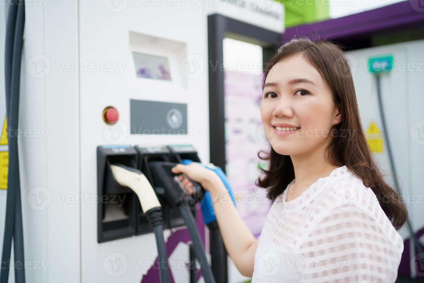 Happy Asian woman holding a DC - CCS type 2 EV charging connector at EV charging station, woman preparing an EV - electric vehicle charging connector for recharge a vehicle. photo