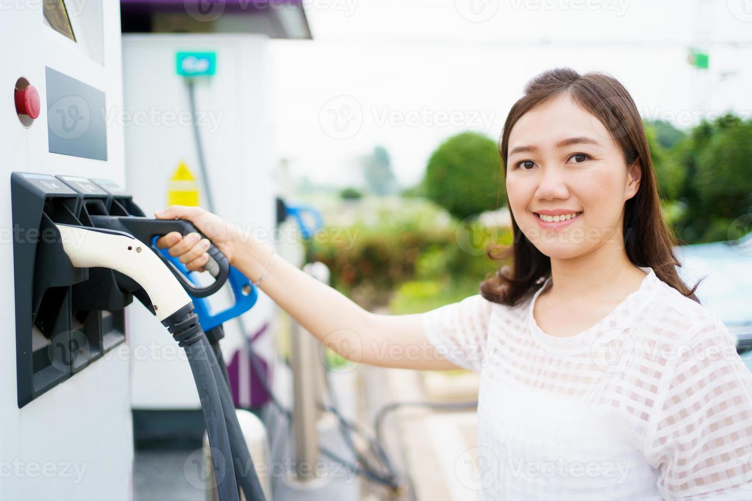 Happy Asian woman holding a DC - CCS type 2 EV charging connector at EV charging station, woman preparing an EV - electric vehicle charging connector for recharge a vehicle. photo
