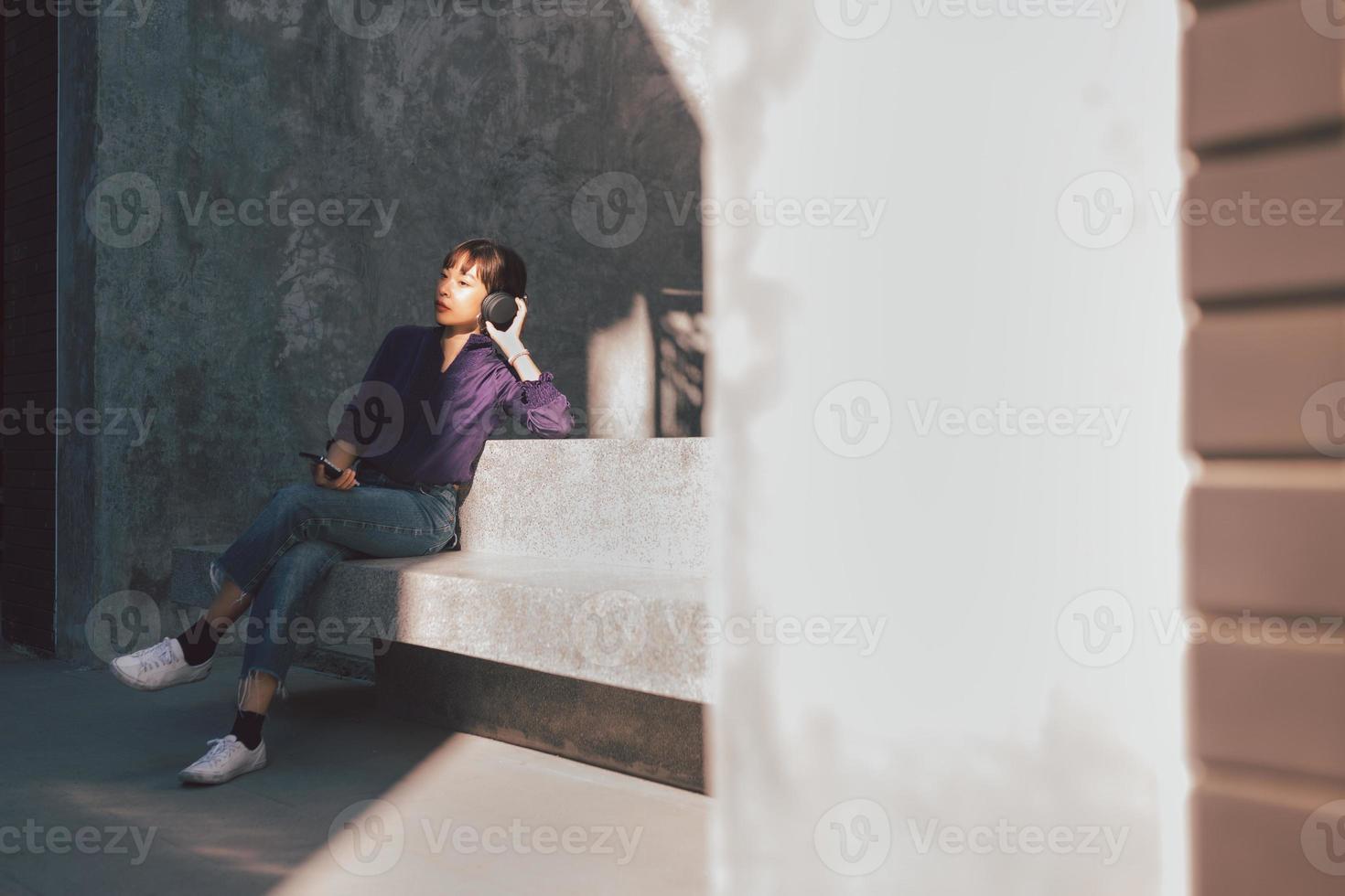 Happy young asian woman listening to music with headphones via smartphone and having fun while sitting side the street photo