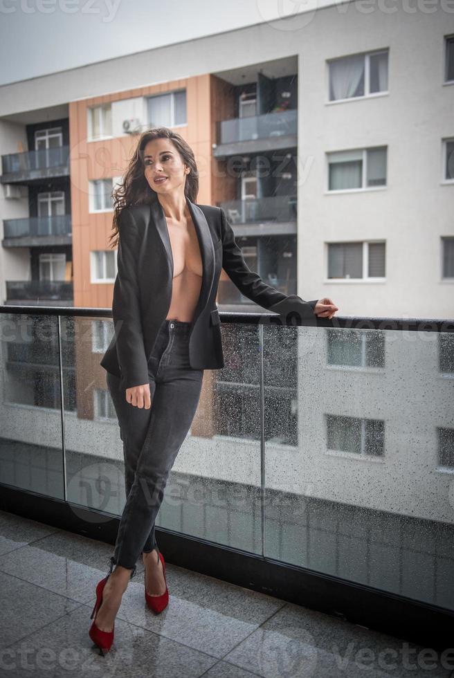 Gorgeous glamour brunette woman with black jacket posing on modern balcony with amazing view on city .Portrait of a Stylish fashionable woman with long legs , black jeans wearing in balcony photo