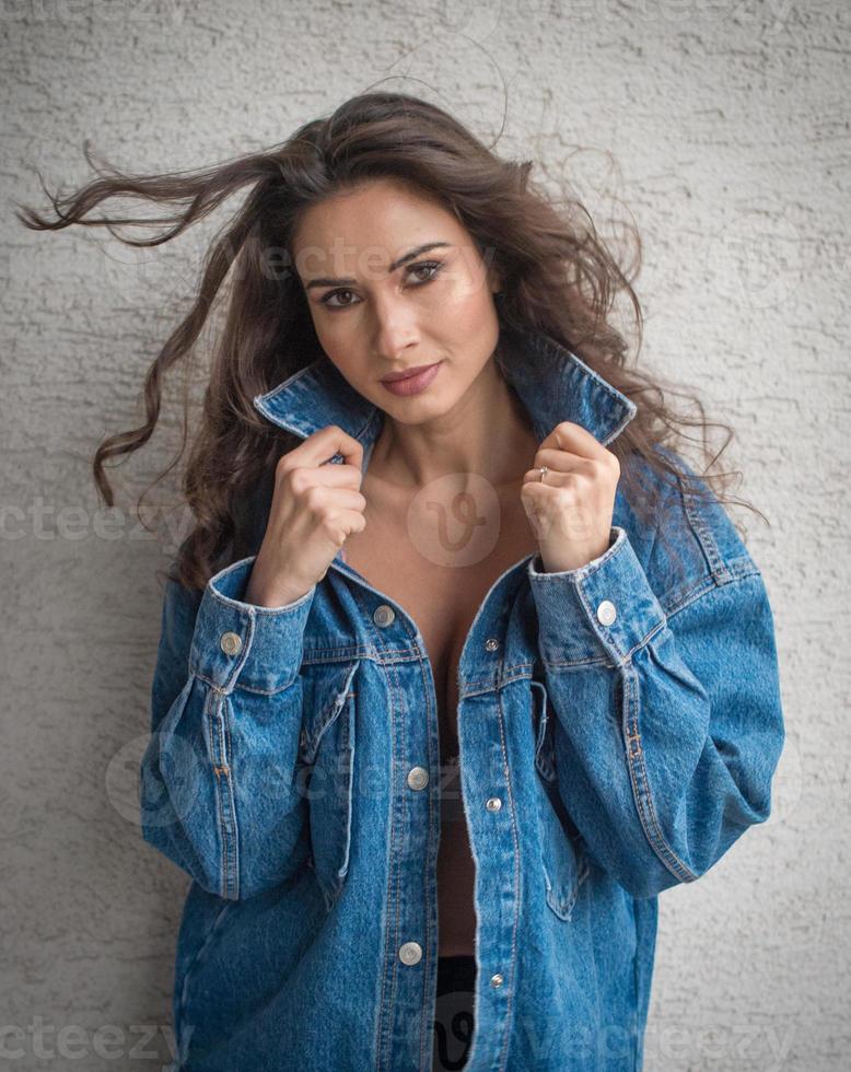 Hermosa mujer morena glamurosa con chaqueta azul posando en un balcón moderno con una vista increíble de la ciudad. Retrato de una elegante mujer de moda con cabello largo, jeans negros en el balcón foto