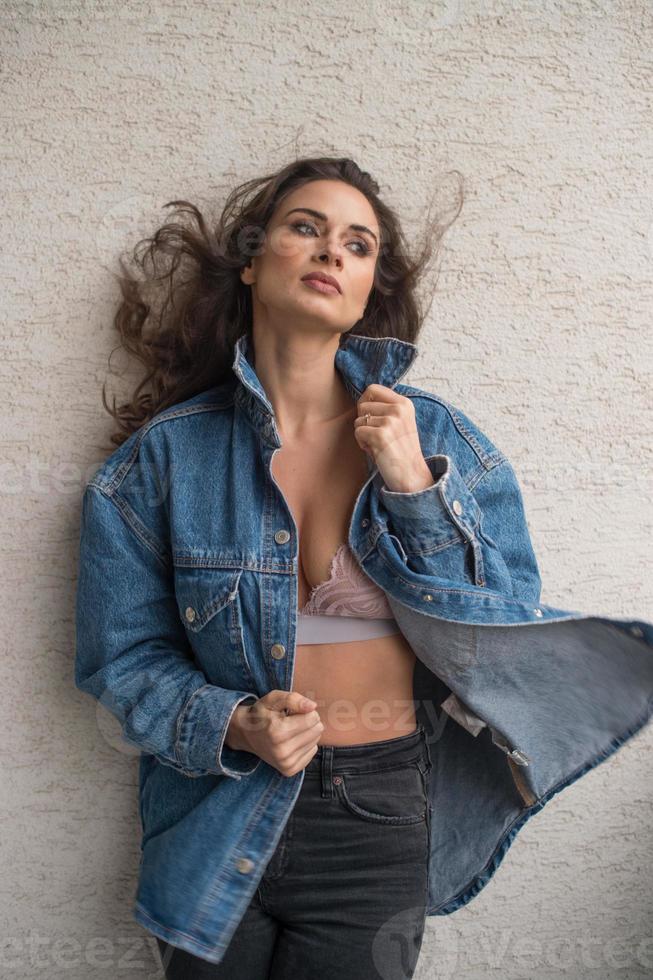 Gorgeous glamour brunette woman with blue  jacket posing on modern balcony with amazing view on city .Portrait of a Stylish fashionable woman with long hair , black jeans wearing in balcony photo