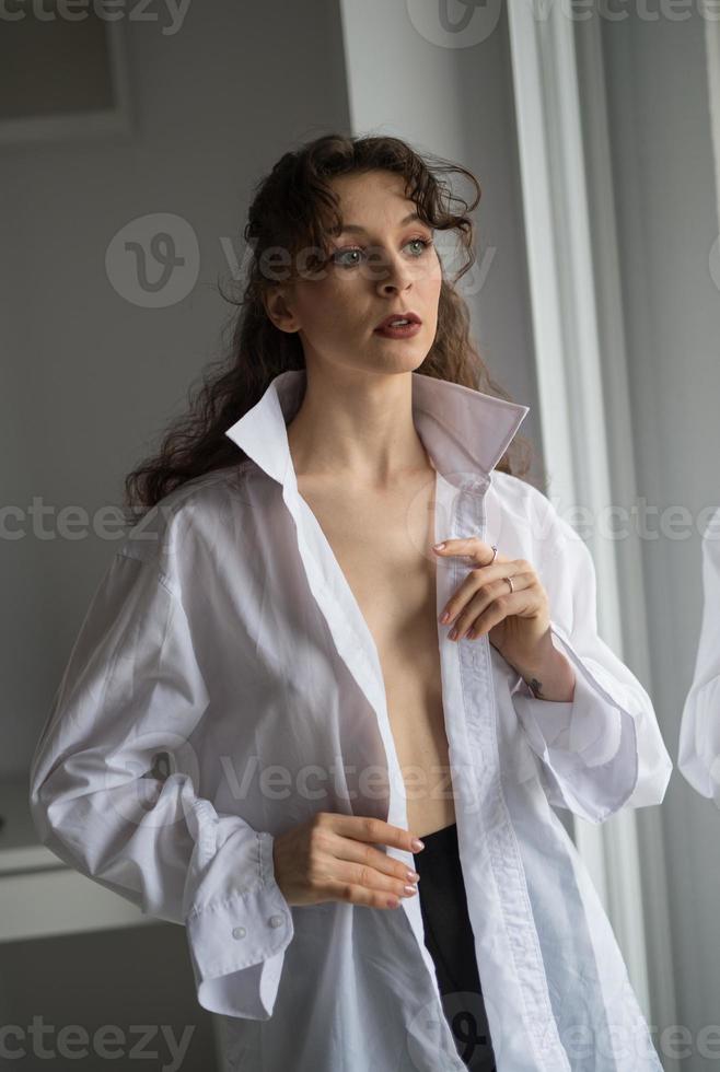 atractiva morena sexy con camisa blanca posando provocativamente, cerca de una ventana, tiro del estudio. retrato de una mujer sensual con el pelo largo, en una clásica escena de boudoir, miró por la ventana foto