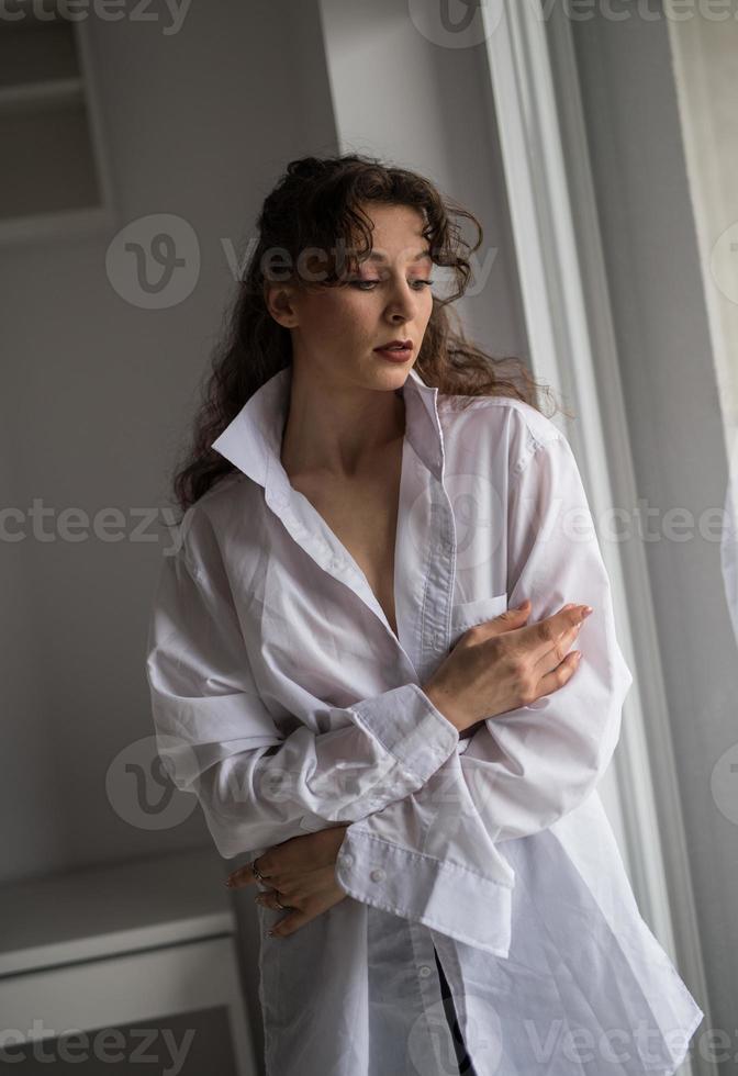 atractiva morena sexy con camisa blanca posando provocativamente, cerca de una ventana, tiro del estudio. retrato de una mujer sensual con el pelo largo, en una clásica escena de boudoir, miró por la ventana foto