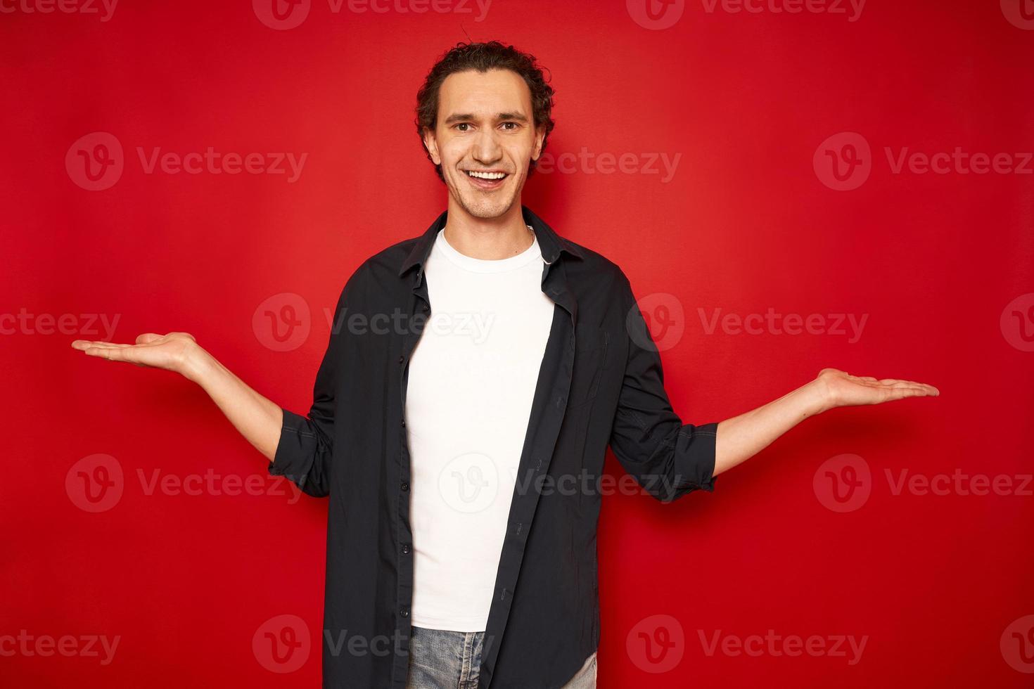 young man holds an empty free space with both hands, offers equal price options. smiling, dressed in casual clothes, isolated on a red background. concept - people, comparison, marketing, advertising photo