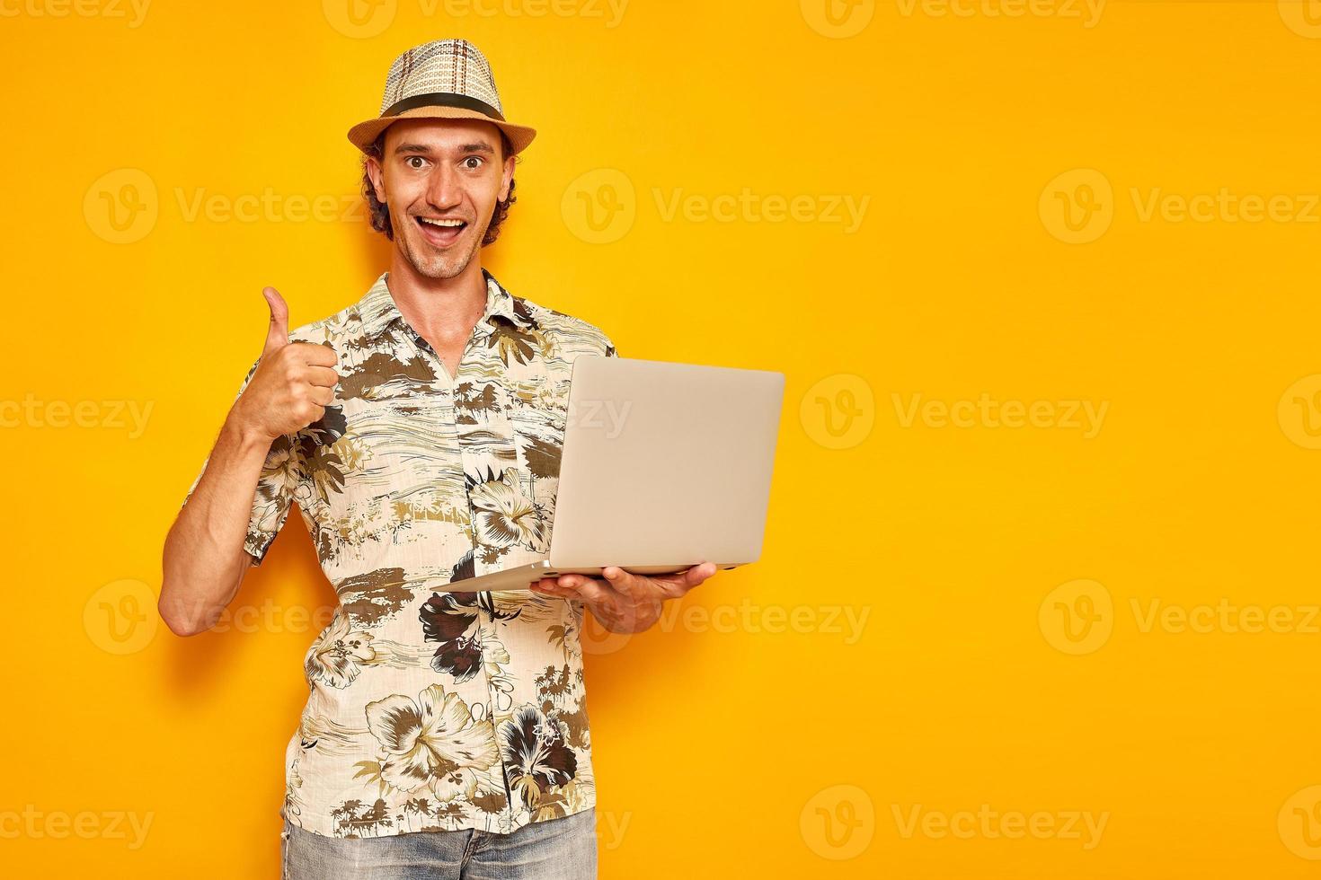 an excited happy male tourist traveler on vacation with laptop in his hand shows thumbs up sign of victory of successful deal, discounts, promotions. isolated on yellow background with space for text. photo