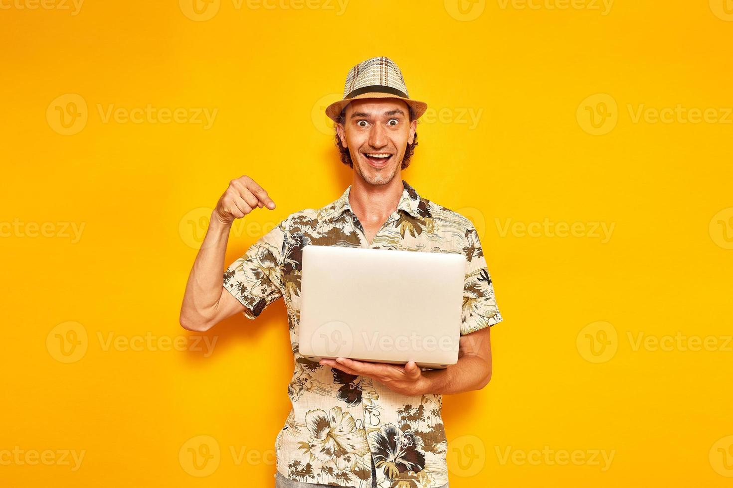 male tourist traveler with laptop in his hands on vacation received message by email with joyful news. he points his finger at device. isolated on yellow background. space for text. concept recreation photo