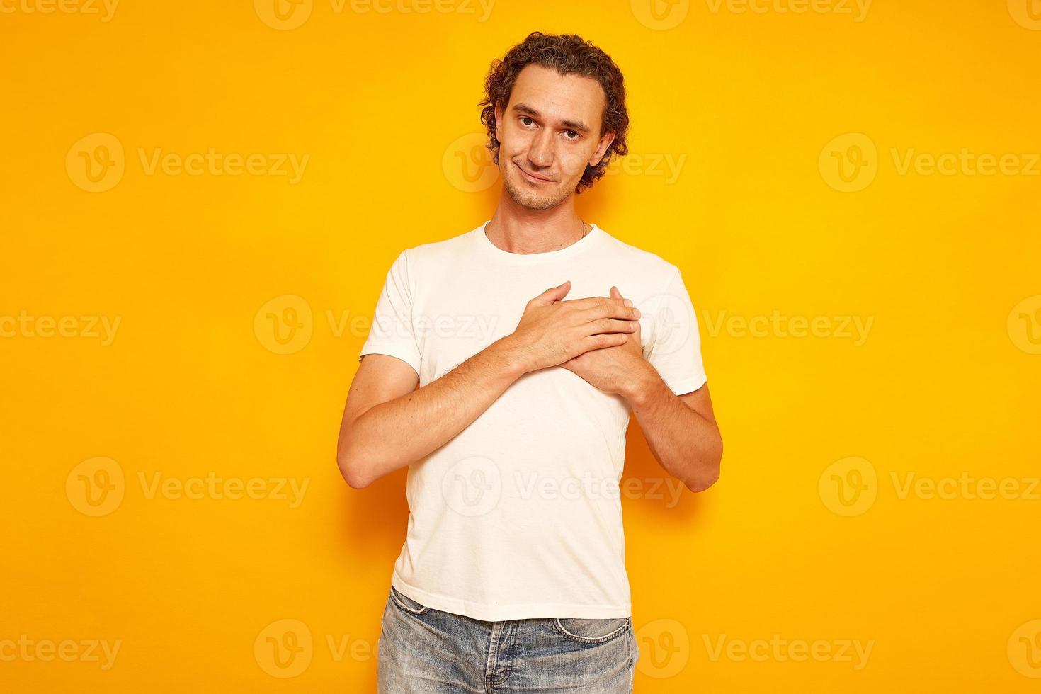 toma interior de un hombre guapo que mantiene las manos en el pecho, es cordial y amigable, expresa gratitud y agradecimiento, tiene una sonrisa agradable, vestido con ropa informal, aislado en la pared amarilla del estudio foto