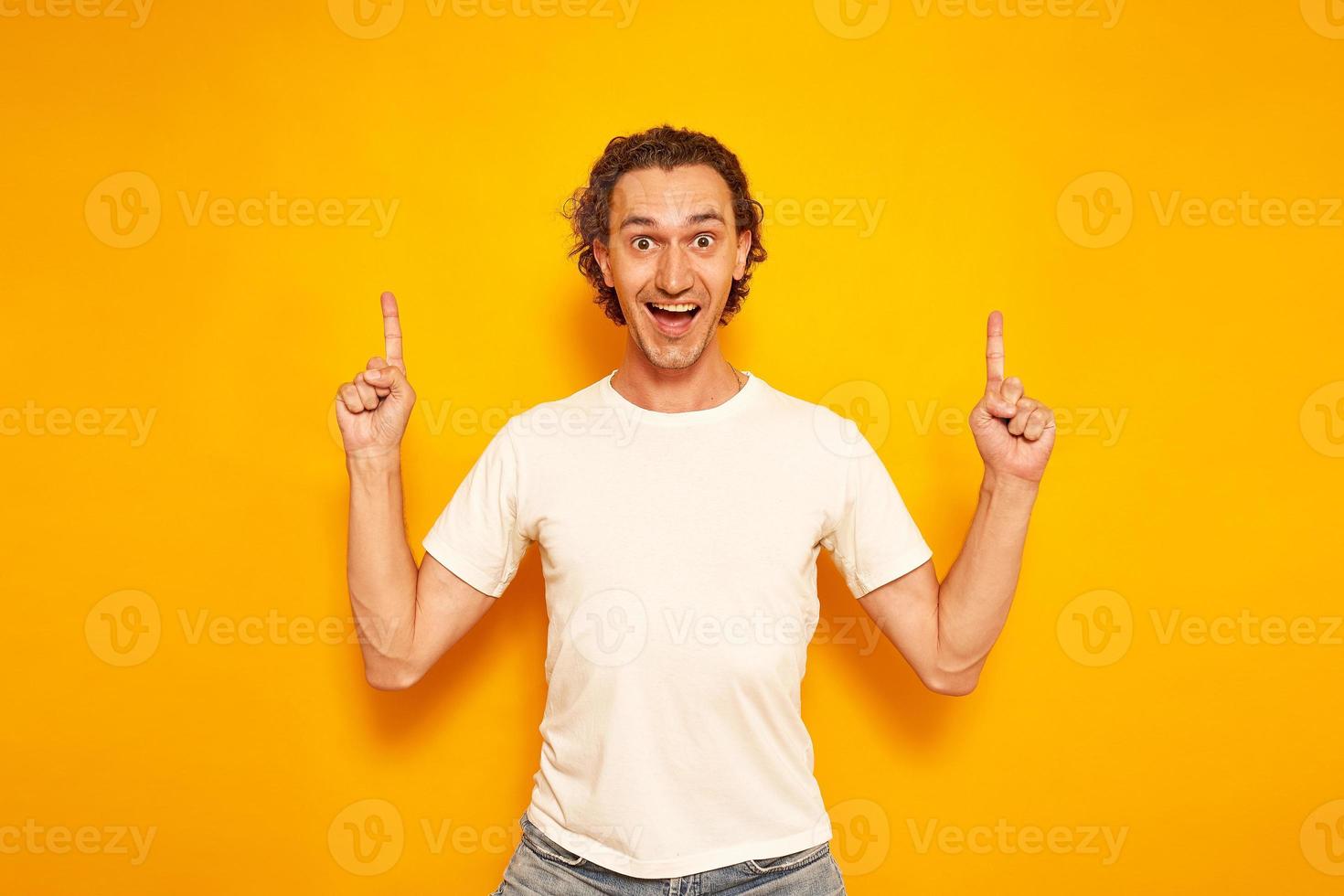 an excited curly-haired man in casual clothes looks forward, points up with his index fingers at an empty area for advertising, attracts attention. isolated on a yellow background with space for text. photo